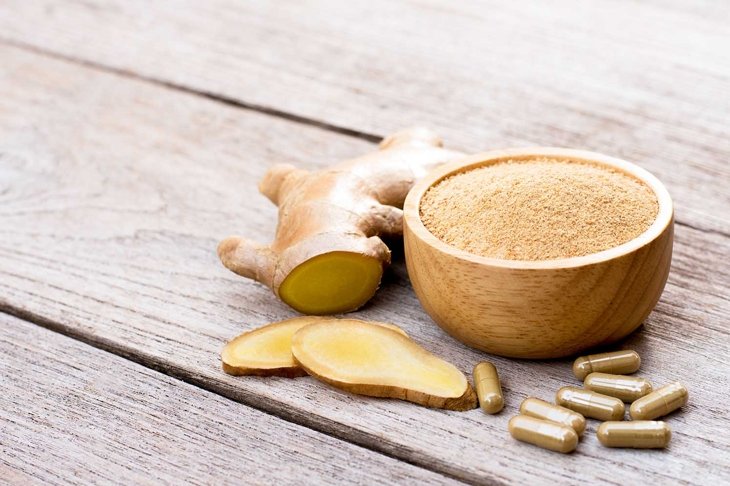Fresh ginger slice and powder capsules with ginger ground in wooden bowl isolated on wood table background. Herbal medicine plant and supplement concept.